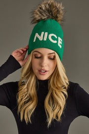 Green beanie with 'NICE' text and fur pom-pom worn by a person against a gray background