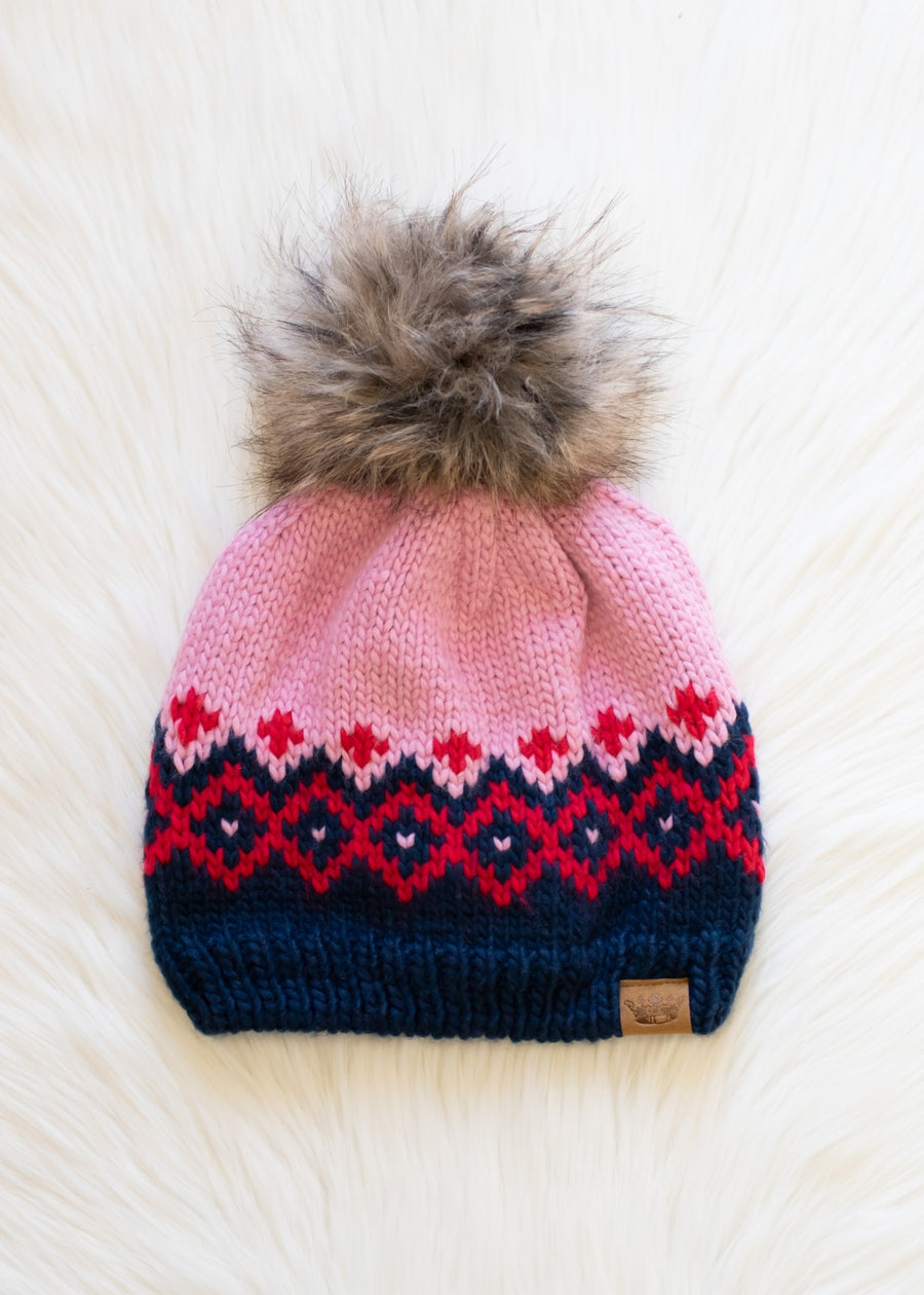 Knit beanie with pink and navy pattern and fur pom-pom on a white background
