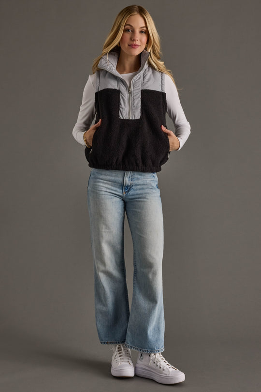 Woman wearing a two-tone vest over a shirt, paired with jeans and white sneakers on a gray background