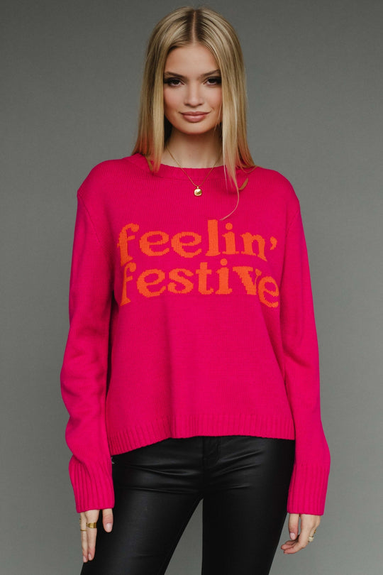 Woman wearing a bright pink sweater with 'feelin' festive' text on a gray background