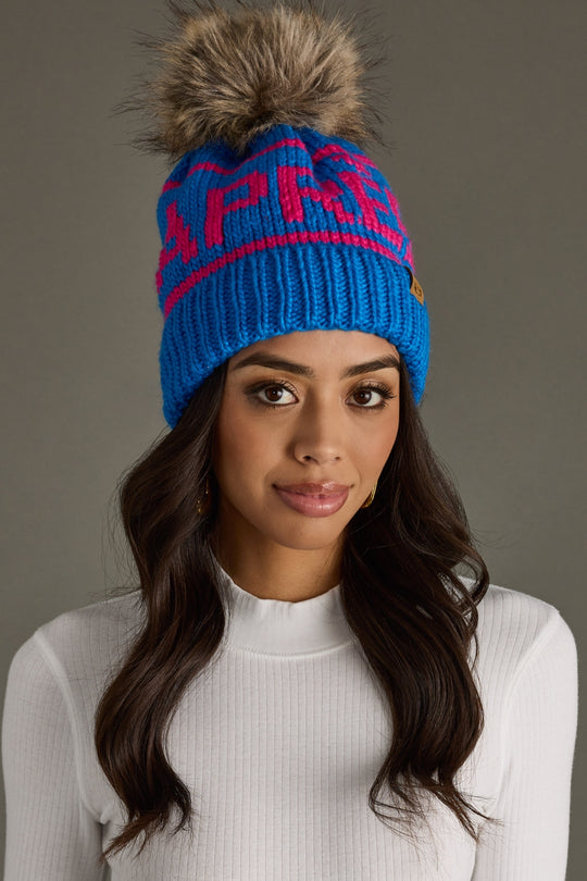 Woman wearing a blue knit beanie with a fur pom-pom on a gray background