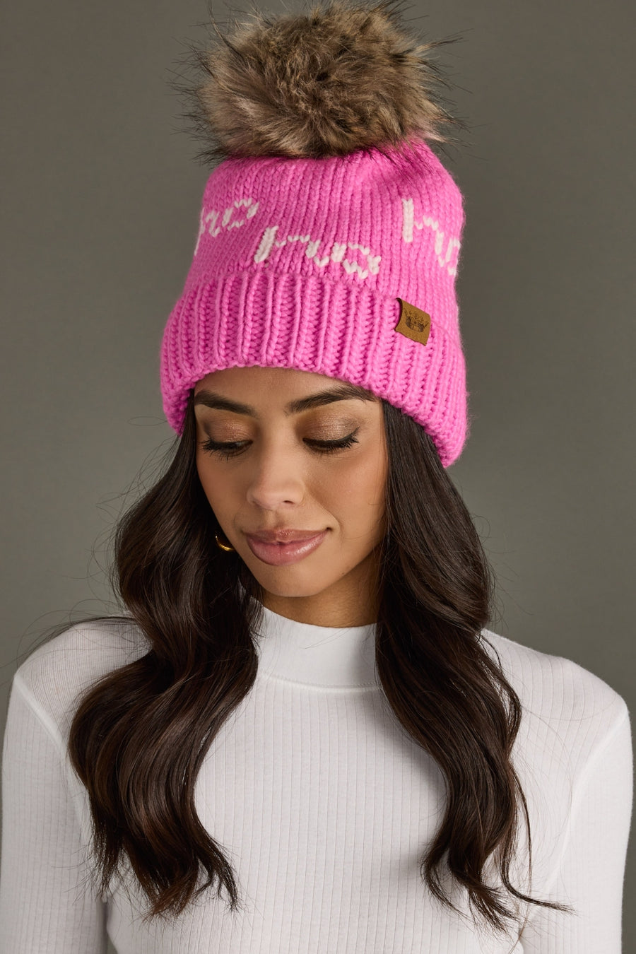 Pink beanie with fur pom-pom worn by a model against a gray background