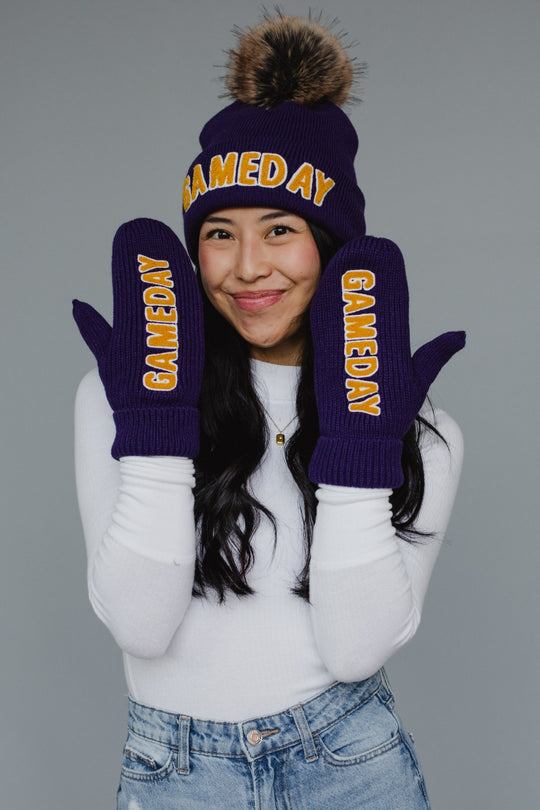 A woman in a white long-sleeve shirt and jeans smiles, wearing Panache Apparel Collections Game Day Mittens - Purple and a matching beanie, both featuring bold yellow GAME DAY lettering.