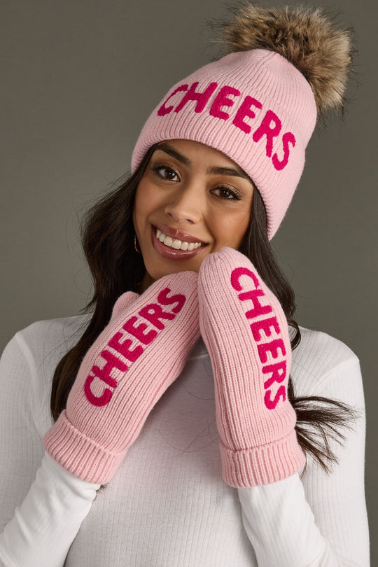 Person wearing pink 'CHEERS' beanie and mittens on a gray background