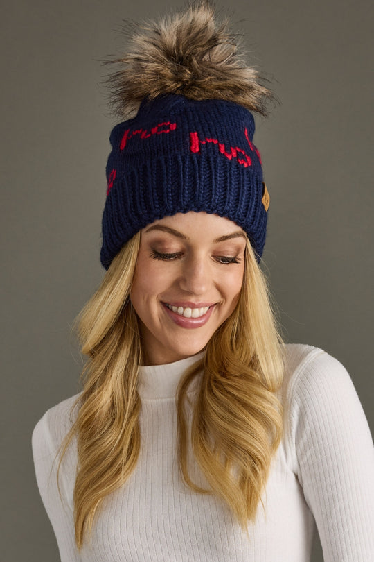 Woman wearing a navy blue beanie with a pom-pom and red text, smiling against a gray background
