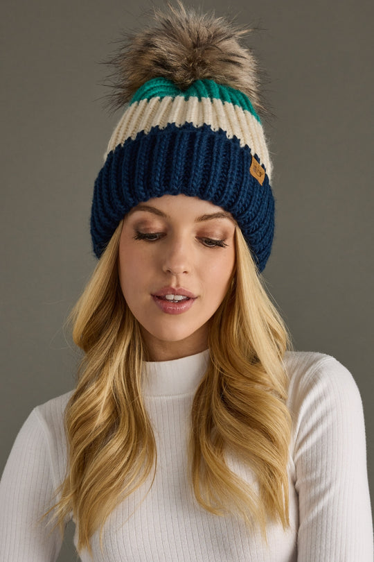 A woman with long blonde hair wears the Panache Apparel Collection So Cozy Pom Hat, featuring navy, cream, and green colorblock stripes with a large faux fur pom-pom, along with a white ribbed turtleneck. She gazes downward with a gentle expression.