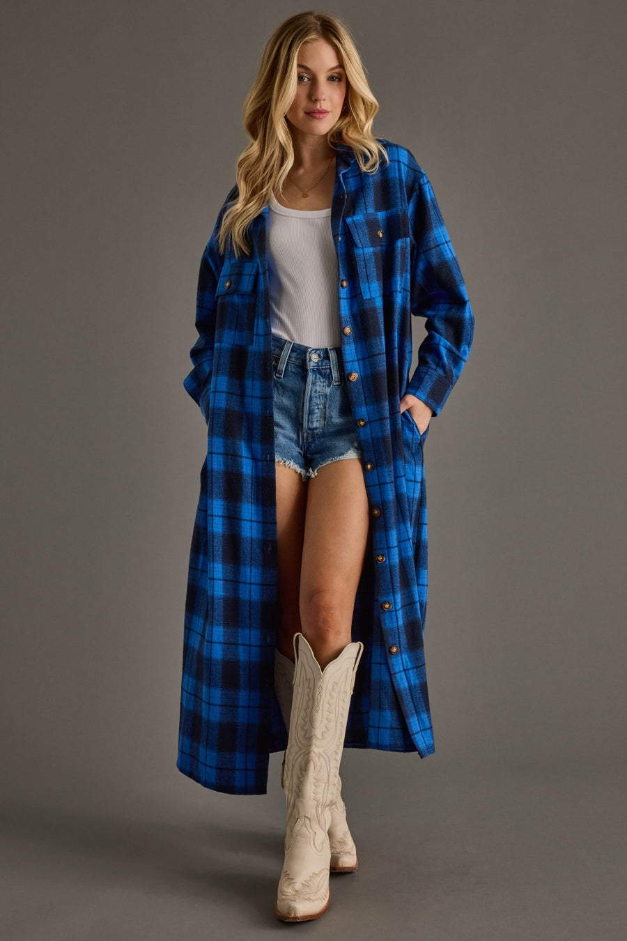 Woman wearing a long blue plaid coat over a white top and denim shorts, standing against a gray background.