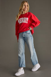 Woman wearing a red sweatshirt with 'Merry' text, blue jeans, and white sneakers on a gray background