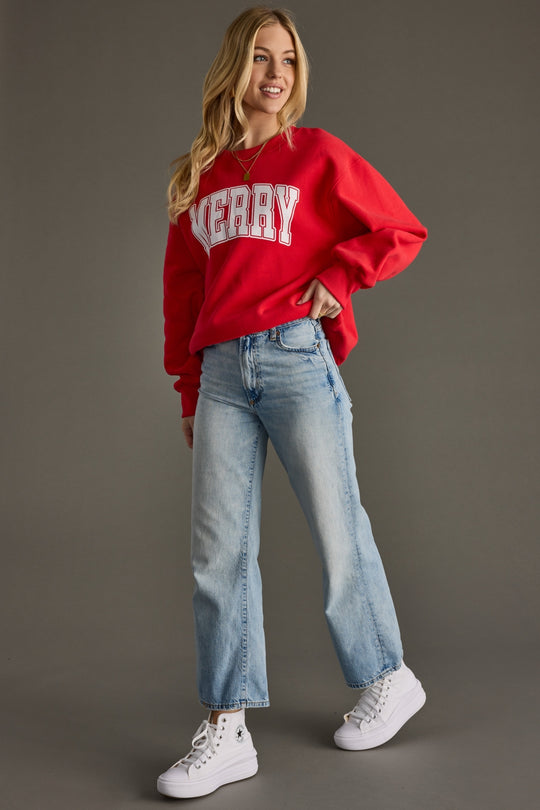 Woman wearing a red sweatshirt with 'Merry' text, blue jeans, and white sneakers on a gray background