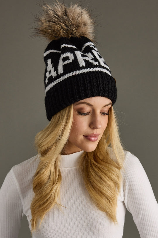 Woman wearing a black beanie with a fur pom-pom and 'APRES' branding against a gray background