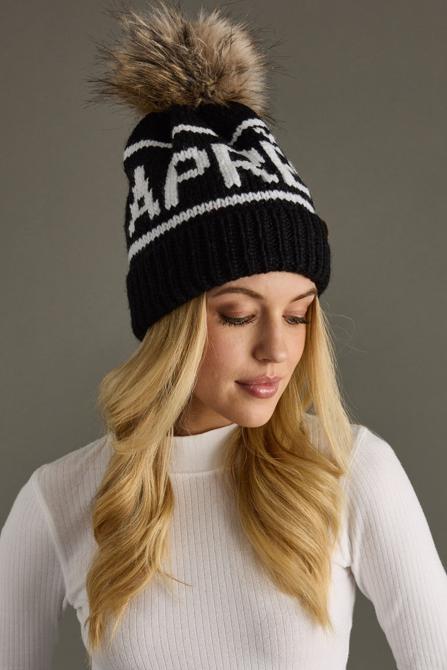 Woman wearing a black beanie with a fur pom-pom and 'APRES' branding against a gray background