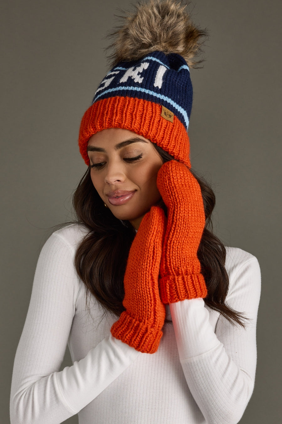 Woman wearing an orange and blue knit hat with a pom-pom and matching scarf against a gray background