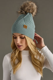 Woman wearing a light blue knit beanie with a fur pom-pom against a gray background