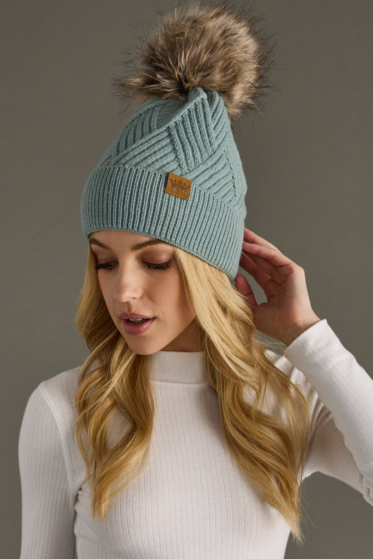 Woman wearing a light blue knit beanie with a fur pom-pom against a gray background