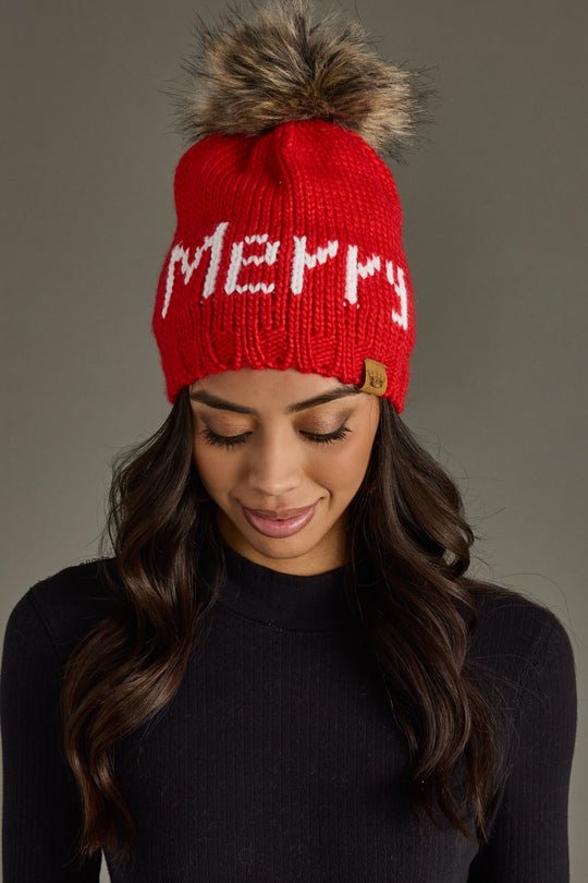 Red knit beanie with 'Merry' text and fur pom-pom worn by a person against a gray background
