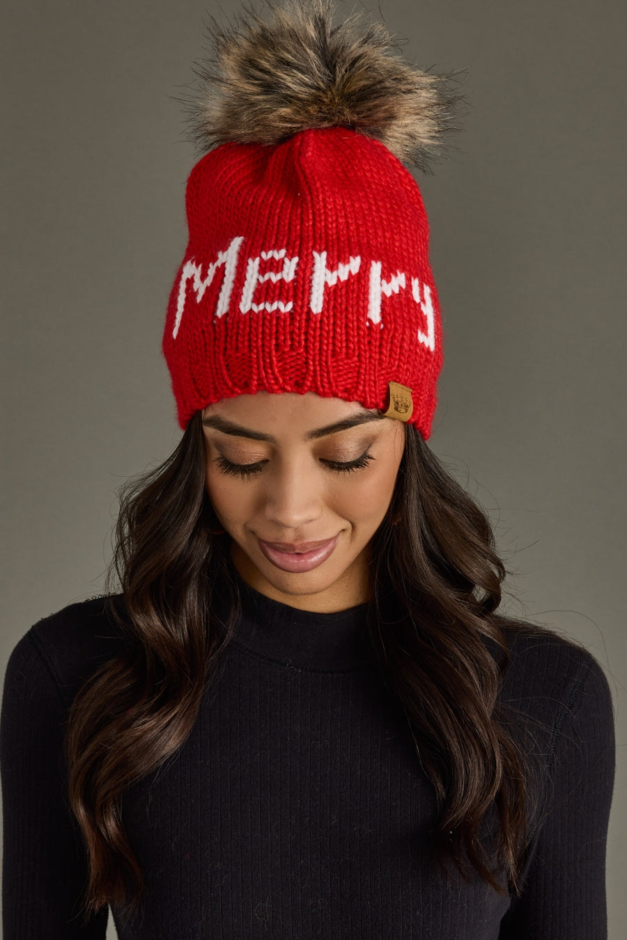 Red knit beanie with 'Merry' text and fur pom-pom worn by a person against a gray background