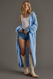 Woman wearing a long light blue cardigan over a white tank top and denim shorts, standing against a gray background.