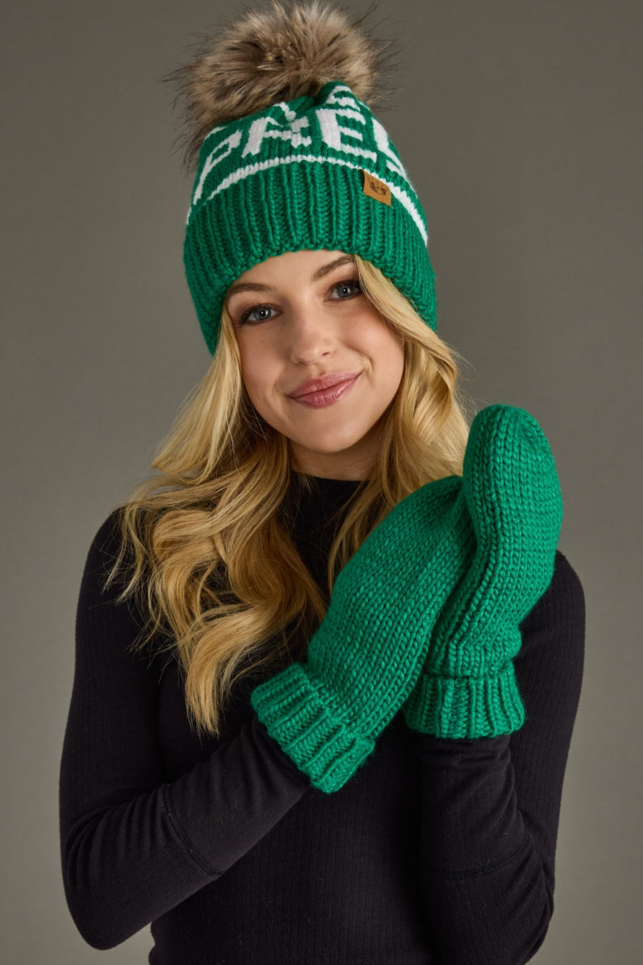 Person wearing a green knit hat and gloves with a brand logo on a gray background