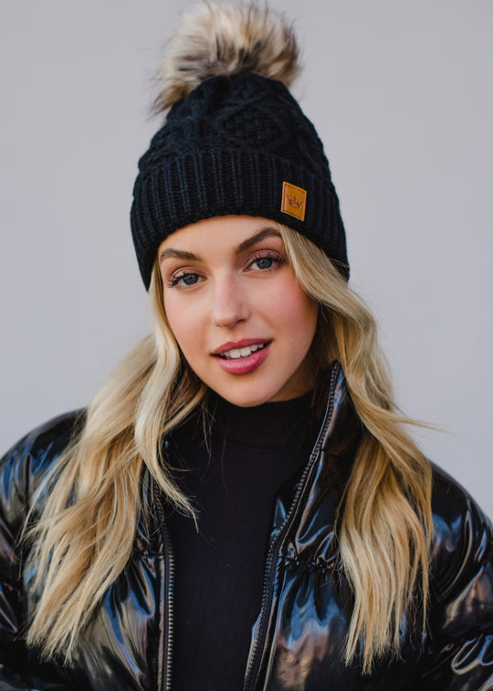 Woman wearing a black knit beanie with a pom-pom against a plain background