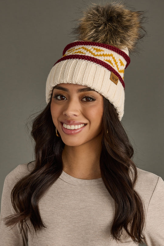 Woman wearing a cream knit beanie with colorful stripes and a pom-pom on a gray background