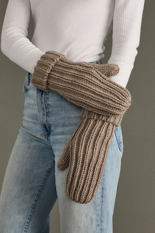 Brown knitted mittens worn by a person with a white long-sleeve shirt and blue jeans on a gray background