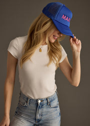 A woman in a fitted white top wears a bright blue baseball cap with bold pink text that reads “MARG.” She’s looking down with a soft smile, arms loosely crossed.