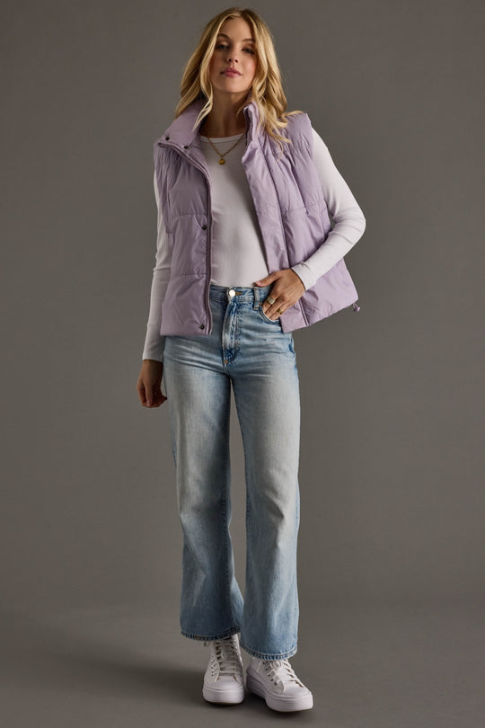 Woman wearing a light purple puffer vest, white shirt, blue jeans, and white sneakers on a gray background