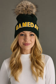 Woman wearing a green beanie with yellow text and a fur pom-pom against a gray background