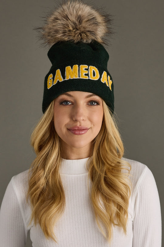 Woman wearing a green beanie with yellow text and a fur pom-pom against a gray background
