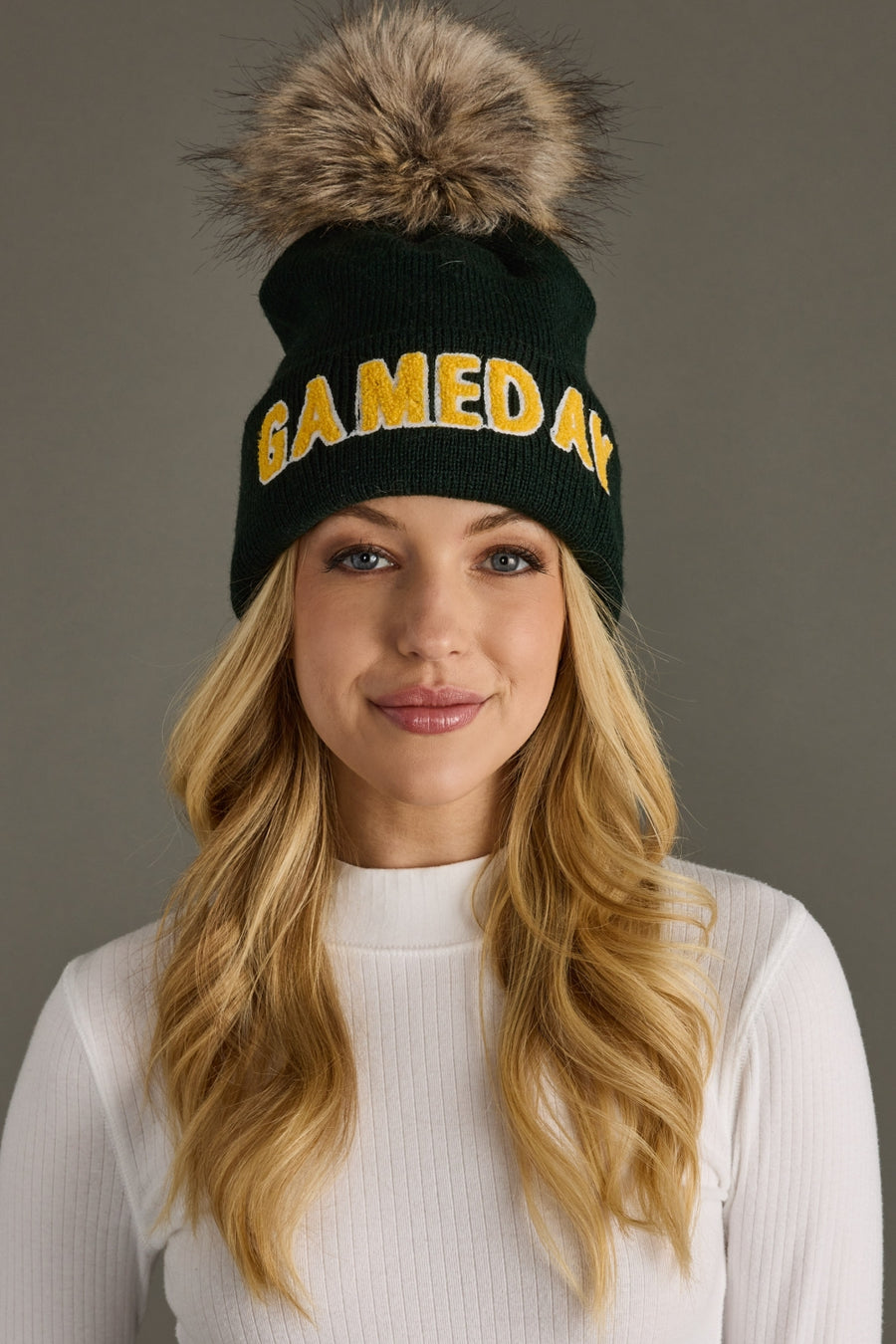 Woman wearing a green beanie with yellow text and a fur pom-pom against a gray background