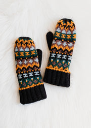 The Valley Creek Mittens by Panache Apparel Collection are acrylic knit mittens featuring colorful orange, green, black, white, and gray patterns, shown on a fluffy white surface.