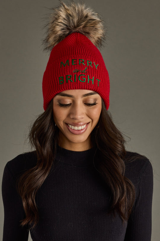 Woman wearing a red knit beanie with 'Merry and Bright' text and a fur pom-pom on a gray background