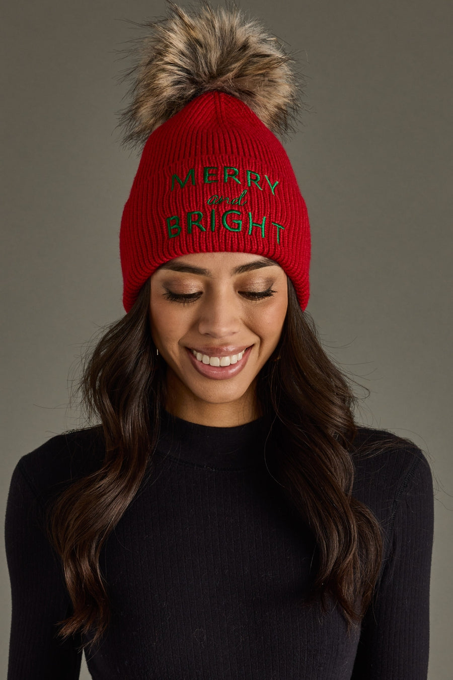 Woman wearing a red knit beanie with 'Merry and Bright' text and a fur pom-pom on a gray background