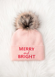 Pink beanie with fur pom-pom and 'Merry and Bright' text on a white background