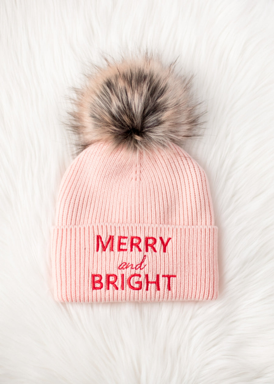 Pink beanie with fur pom-pom and 'Merry and Bright' text on a white background