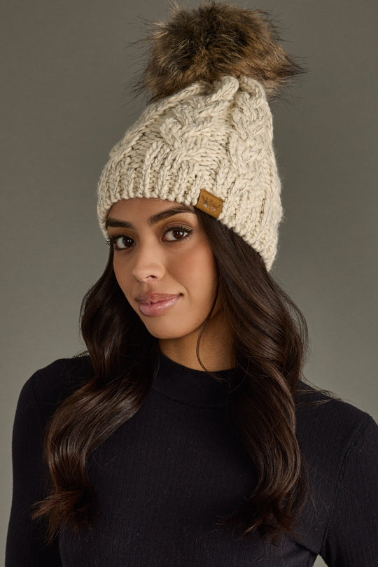 Woman wearing a beige knitted beanie with a fur pom-pom against a gray background