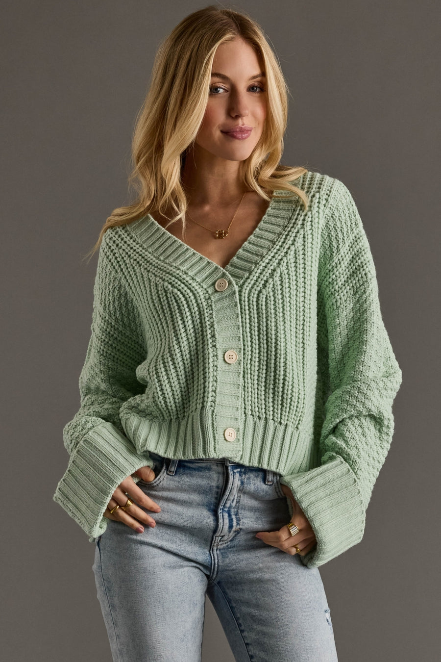 Woman wearing a light green knitted cardigan and blue jeans against a gray background