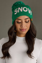 A woman wears the Panache Apparel Collection Snow Pom Hat - Green, featuring a playful yarn pom and SNOW text, paired with a white ribbed top and gold hoop earrings as she looks down and smiles softly.