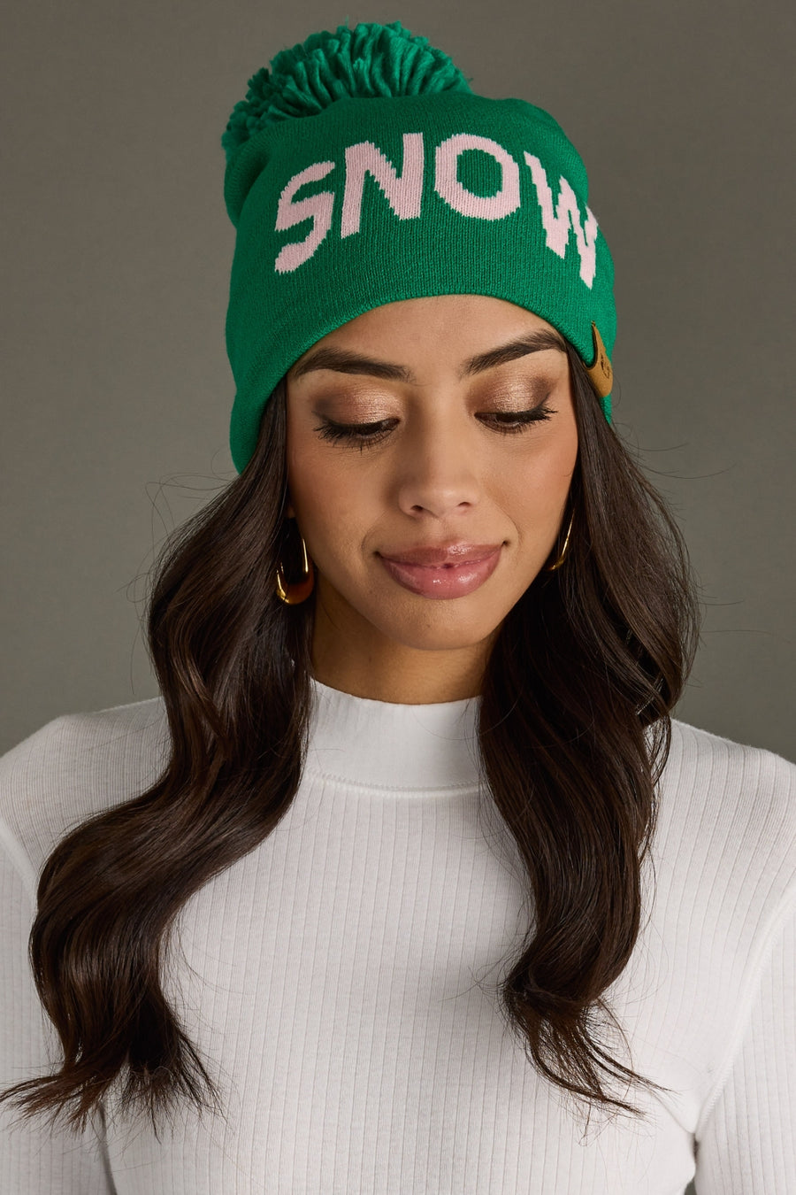 A woman wears the Panache Apparel Collection Snow Pom Hat - Green, featuring a playful yarn pom and SNOW text, paired with a white ribbed top and gold hoop earrings as she looks down and smiles softly.