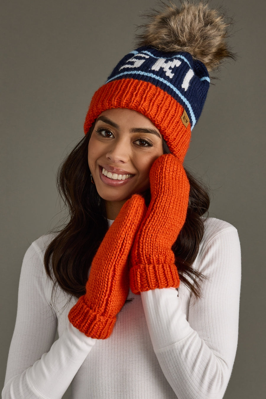 Woman wearing an orange and blue knit hat with a fur pom-pom and matching gloves against a gray background