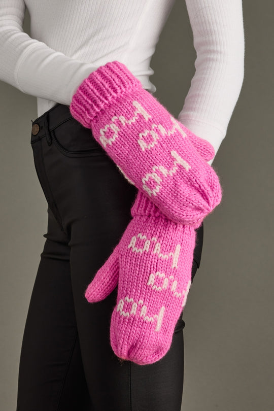 Pink knitted mittens with text held by a person wearing a white long-sleeve shirt and black pants.