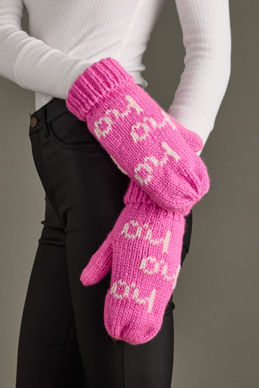 Pink knitted mittens with text held by a person wearing a white long-sleeve shirt and black pants.