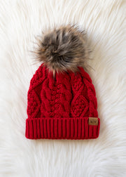 The Red Cable Knit Pom Hat from Panache Apparel Collection rests on a soft, white, fluffy surface. This hat showcases a classic cable knit pattern in vibrant red, features a cozy fleece lining for added warmth, and is topped with a brown faux fur pom. A small brown tag accents the folded brim.