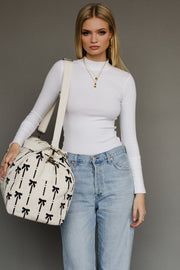 Woman wearing a white long-sleeve top and blue jeans, holding a bag with black bow pattern.