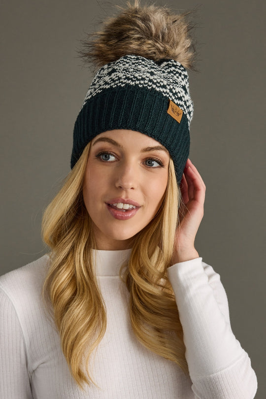 Woman wearing a dark green knit beanie with a fur pom-pom on a brown background