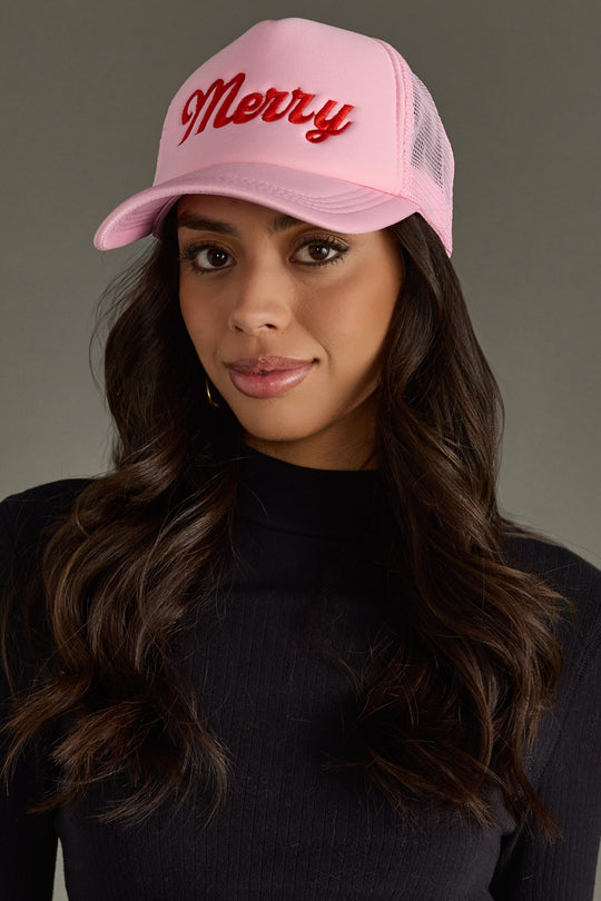 Woman wearing a pink trucker hat with 'Merry' text on a gray background