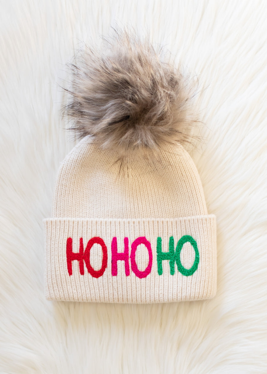 The Ho Ho Ho Pom Hat by Panache Apparel Collection is a beige knit hat with a large faux fur pom-pom and “HO HO HO” embroidered in red, pink, and green on the cuff. Cozy fleece lined for warmth.