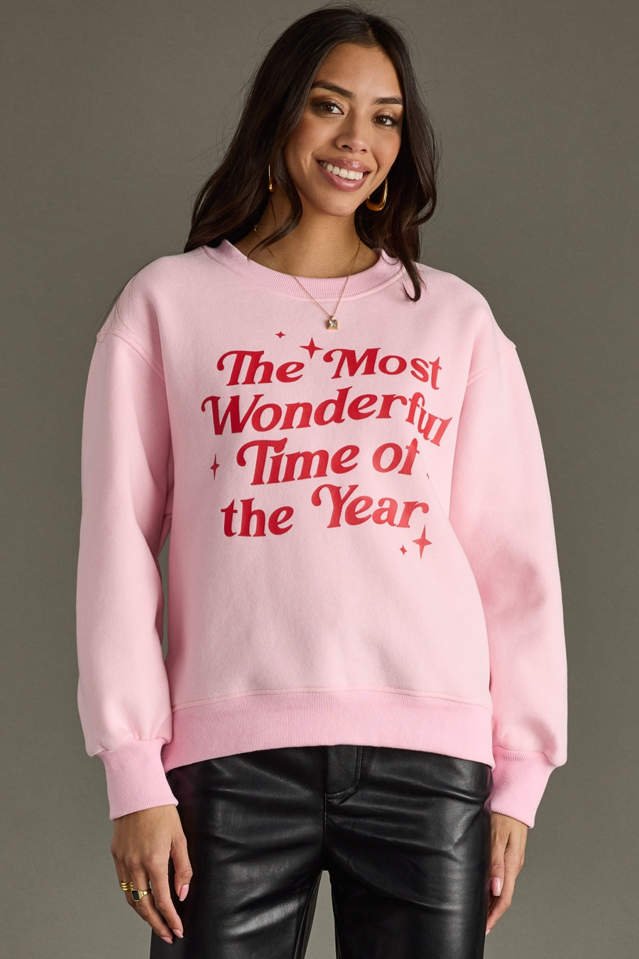 A woman smiles against a gray background wearing a pink oversized Panache Apparel Collection "The Most Wonderful Time Of The Year Sweatshirt" with black leather pants.