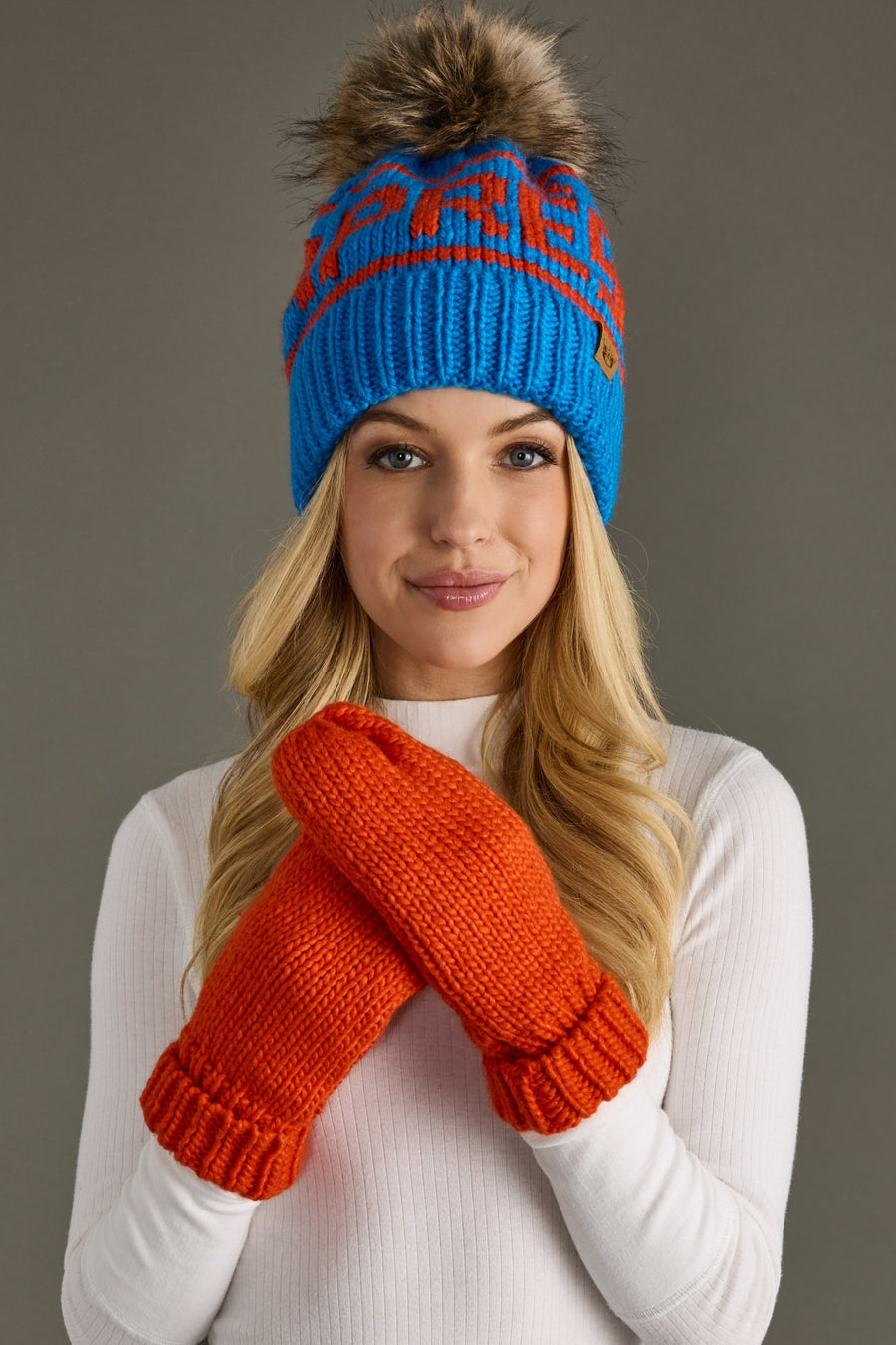 Woman wearing a blue knit hat with a pom-pom and orange mittens against a gray background
