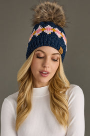 Woman wearing a blue knit beanie with a colorful pattern and fur pom-pom on a gray background